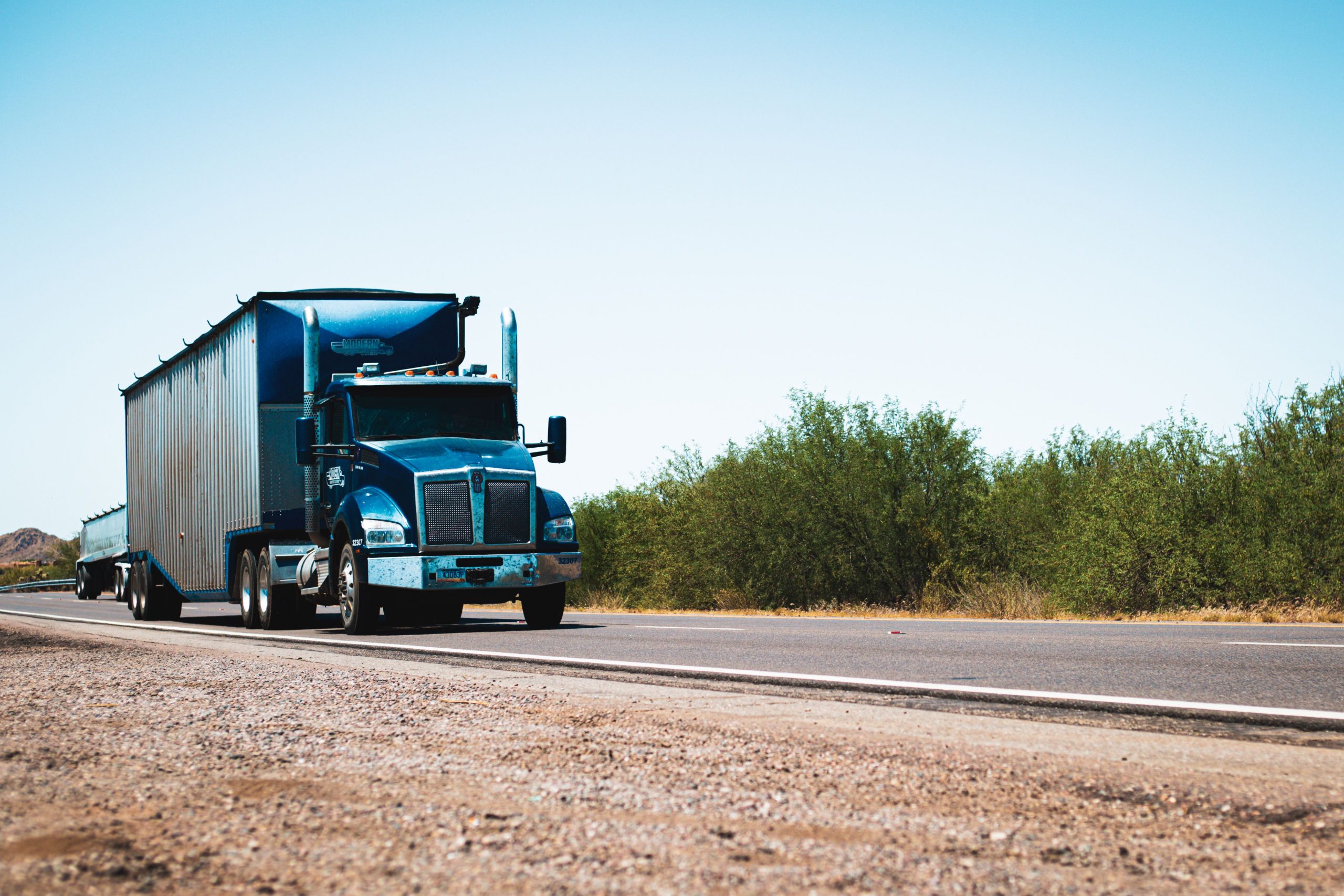 semi-truck-breakdown-far-from-home Blue semi truck traveling alone on a rural highway under a clear blue sky, far from home
