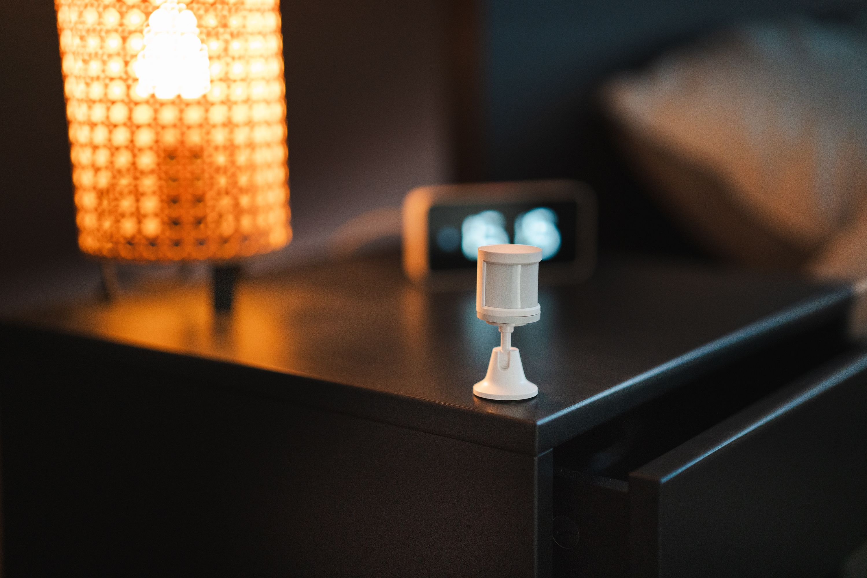 Bedroom with a smart motion sensor, lamp, and alarm clock on a nightstand.